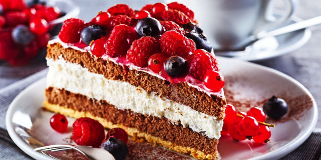 Slice of gourmet fresh berry cake on a plate with layered cream topped with fresh autumn raspberries, blueberries and redcurrants in a close up view on a plate